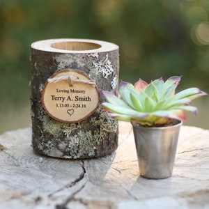 May include: A wooden planter with a bark-like texture and a small wooden plaque that reads "In Loving Memory Terry A. Smith 1.13.03 - 2.24.18" with a heart symbol. A small succulent plant is in a silver pot next to the planter.