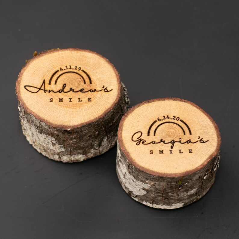May include: Two wooden slices with engraved text and rainbow designs. The top slice reads "Andrew's Smile" with the date "4.11.19". The bottom slice reads "Georgia's Smile" with the date "6.24.20".