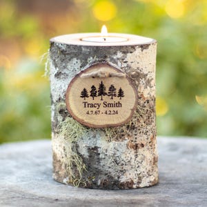 May include: A cylindrical birch log candle holder with a rough, textured bark exterior and light green lichen accents. A small, circular wooden plaque is attached to the front, featuring a printed forest of five pine trees, the name Tracy Smith, and the dates 4.7.67 - 4.2.24.