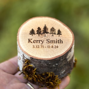 May include: A personalized wooden ring box made from a tree trunk slice. The top features an engraved forest scene, the name "Kerry Smith", and dates. The sides of the box are bark, with moss accents.
