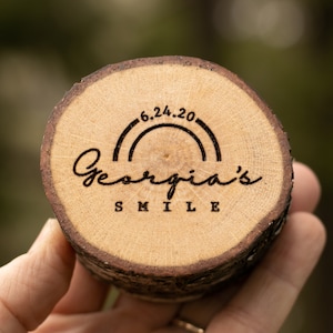 May include: A wooden round disc with a natural wood grain finish. The disc has a rainbow arc with the date "6.24.20" above the text "Georgia's Smile".