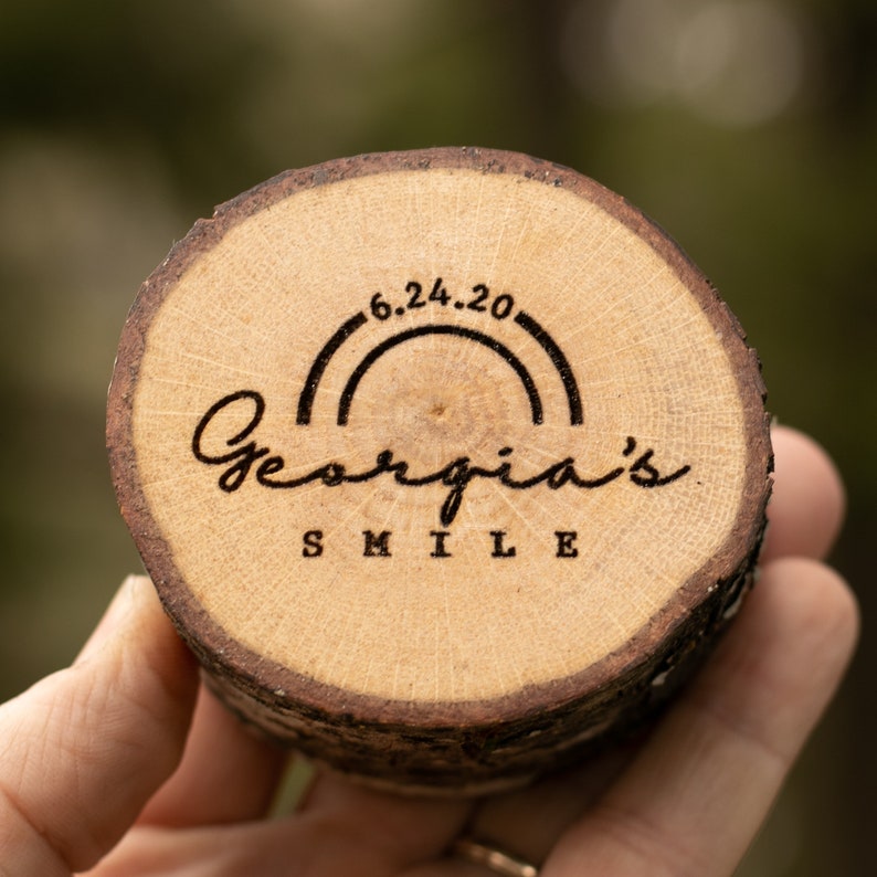 May include: A wooden round disc with a natural wood grain finish. The disc has a rainbow arc with the date "6.24.20" above the text "Georgia's Smile".