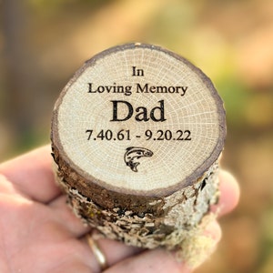May include: A circular wooden log slice with bark edges, featuring laser-engraved text that reads In Loving Memory Dad 7.40.61 - 9.20.22, accompanied by a small graphic of a jumping fish. The wood grain is visible on the flat top surface.