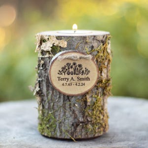 May include: A wooden candle holder with a bark texture and moss accents. It features a small wooden plaque with the text "Terry A. Smith 4.7.63 - 4.2.24" and a floral design.