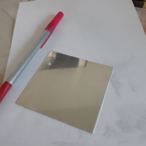 May include: A silver square mirror on a white surface with a red pen.