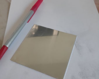 May include: A silver square mirror on a white surface with a red pen.