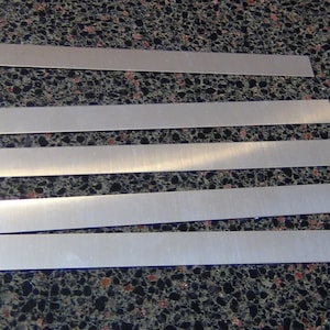May include: Five silver metal strips, all the same size and shape, are laid out on a dark gray speckled surface.