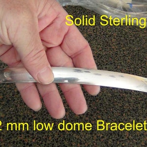 May include: A close-up of a silver bracelet band, 15 mm wide and 2 mm thick, with a low dome shape. The band is made of solid sterling silver. "15 x 2 mm low dome Bracelet band" is written in yellow text at the bottom of the image. "Solid Sterling Silver" is written in yellow text at the top of the image.