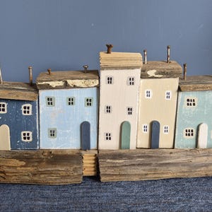 May include: A wooden village scene with five houses in pastel shades. Each house has small windows, doors, and metal chimney details. The houses are mounted on driftwood, giving a rustic, weathered appearance. The houses are painted in blue, cream, and green.