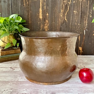 May include: A large, round, hammered copper planter with a wide, flared rim and a bulbous body. The metal surface features a textured, dimpled finish that reflects light. A single, shiny red apple sits on the surface next to the copper vessel.