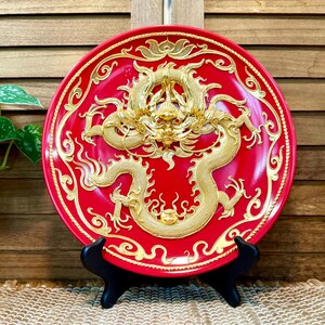 May include: A red decorative plate with a gold dragon design. The dragon is intricately detailed with a raised relief, and the plate is supported by a black stand. The plate has a diameter of approximately 10 inches.