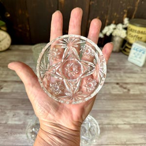 May include: A clear, circular glass coaster featuring a pressed starburst pattern with a central star motif surrounded by four smaller star designs and radiating geometric lines, creating a textured, reflective surface across the entire piece.