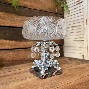 May include: A crystal bowl with a cut glass design sits on a silver stand with crystal drops hanging from the base. The stand rests on a black marble base.