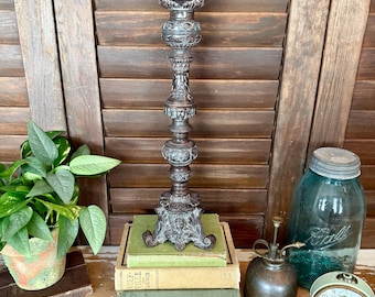 May include: Ornate, dark metal candlestick holder with detailed carvings. The candlestick is tall and stands on a decorative base. It is placed on top of a stack of books. The background includes a plant, a glass jar, and a vintage clock.