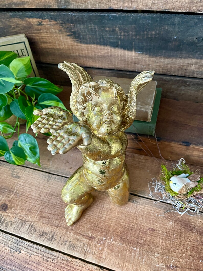 Puede incluir: Una figurita de querub&iacute;n dorada con los brazos y las alas extendidos. El querub&iacute;n est&aacute; de pie sobre una superficie de madera.