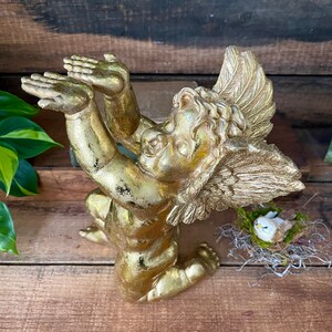 Puede incluir: Una figurita de querub&iacute;n de oro con los brazos y las alas extendidos, arrodillada sobre una superficie de madera. El querub&iacute;n tiene una cara delicada y detallada y est&aacute; adornada con pan de oro.