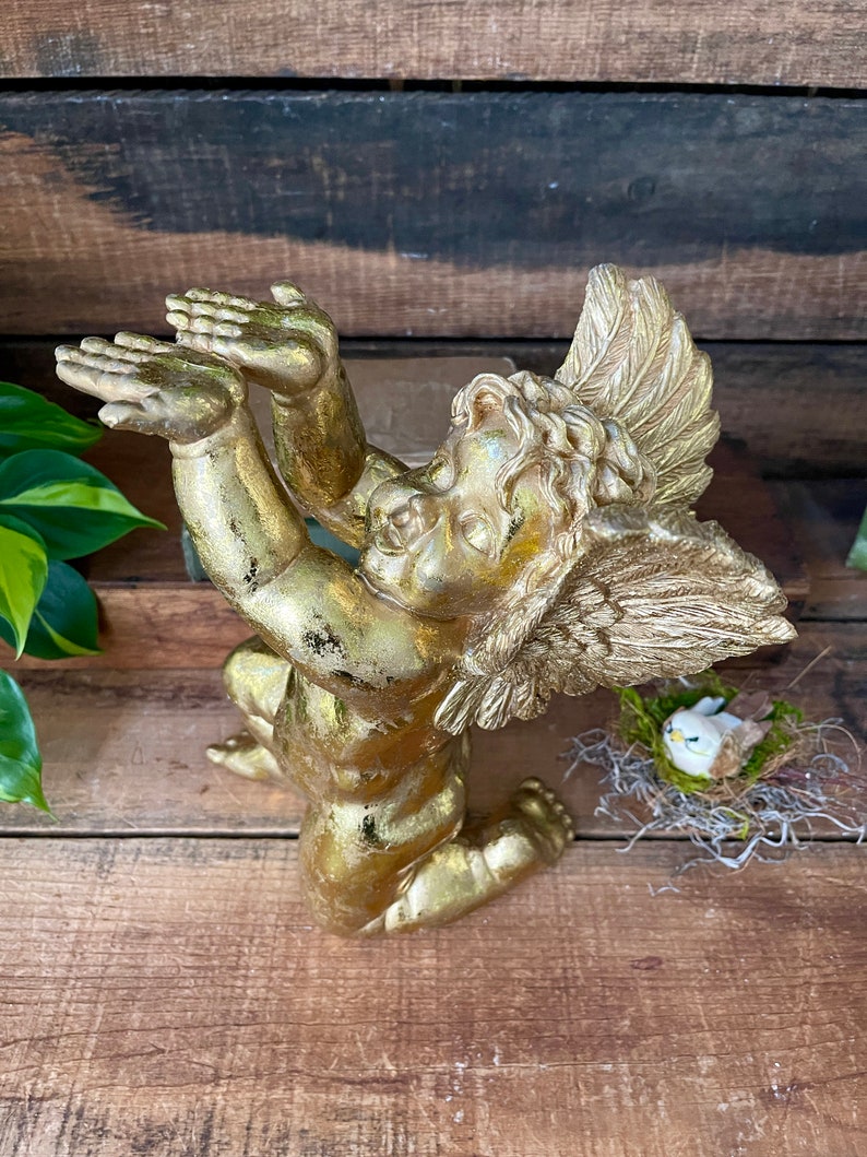 Puede incluir: Una figurita de querub&iacute;n de oro con los brazos y las alas extendidos, arrodillada sobre una superficie de madera. El querub&iacute;n tiene una cara delicada y detallada y est&aacute; adornada con pan de oro.