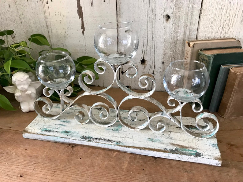 Op de afbeelding: Witte metalen kaarsenhouder met een verweerde afwerking en drie glazen kaarsenhouders. De kaarsenhouder staat op een houten basis.