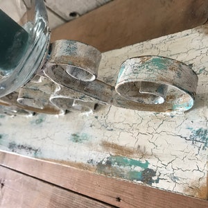 Op de afbeelding: Een wit houten plank met een verweerde afwerking, met een metalen kaarsenhouder met een scroll-ontwerp. De kaarsenhouder is geschilderd in een wit en blauw kleurenschema, met een verweerde uitstraling.