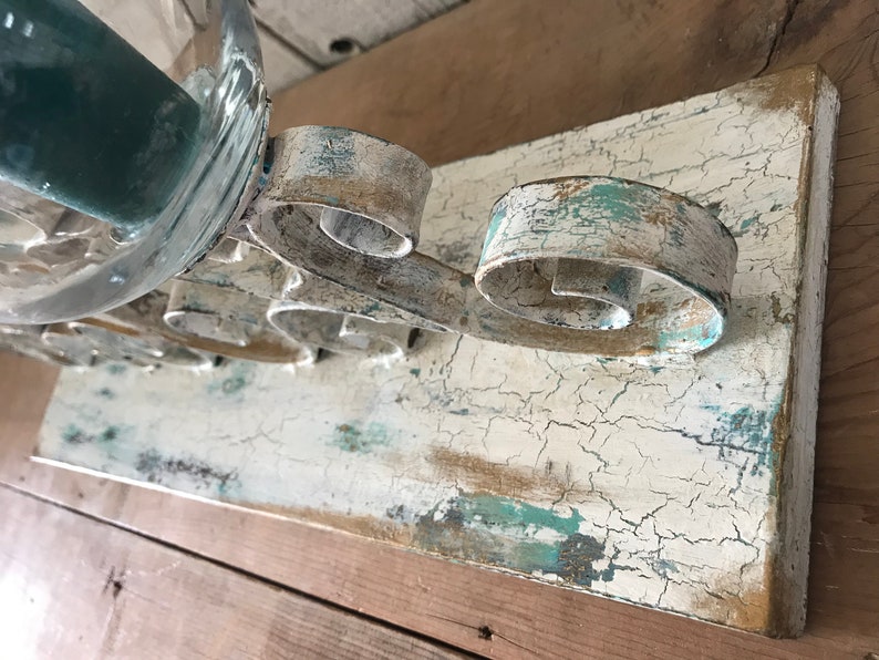 Op de afbeelding: Een wit houten plank met een verweerde afwerking, met een metalen kaarsenhouder met een scroll-ontwerp. De kaarsenhouder is geschilderd in een wit en blauw kleurenschema, met een verweerde uitstraling.