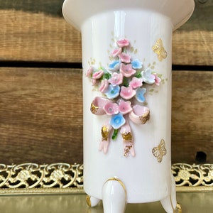Peut inclure: Un vase en porcelaine blanche avec un motif floral et des accents dor&eacute;s. Le vase a trois pieds et une large ouverture.