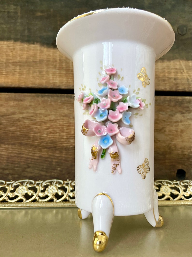 Peut inclure: Un vase en porcelaine blanche avec un motif floral et des accents dor&eacute;s. Le vase a trois pieds et une large ouverture.