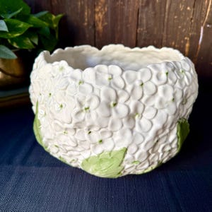 May include: A white ceramic bowl featuring a three-dimensional relief pattern of hydrangea flowers covering the entire exterior surface, accented with small green leaves at the base and subtle green centers on the individual petals.
