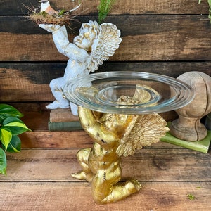 Puede incluir: Una estatua de querub&iacute;n dorada sosteniendo un soporte para pastel de vidrio transparente. El querub&iacute;n tiene alas y est&aacute; arrodillado sobre una superficie de madera.