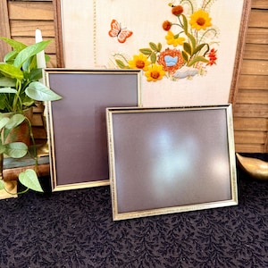 May include: Two rectangular picture frames with thin gold-toned metal borders and dark gray backing boards. One frame is oriented vertically and the other horizontally. Behind them sits a framed piece of fabric featuring embroidered flowers and a butterfly.