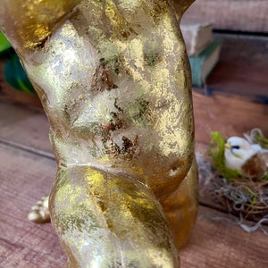 Puede incluir: Estatua de querub&iacute;n de color dorado con un acabado desgastado. La estatua est&aacute; en posici&oacute;n de pie con una pierna levantada y la otra doblada. El querub&iacute;n sostiene un objeto peque&ntilde;o en sus manos.