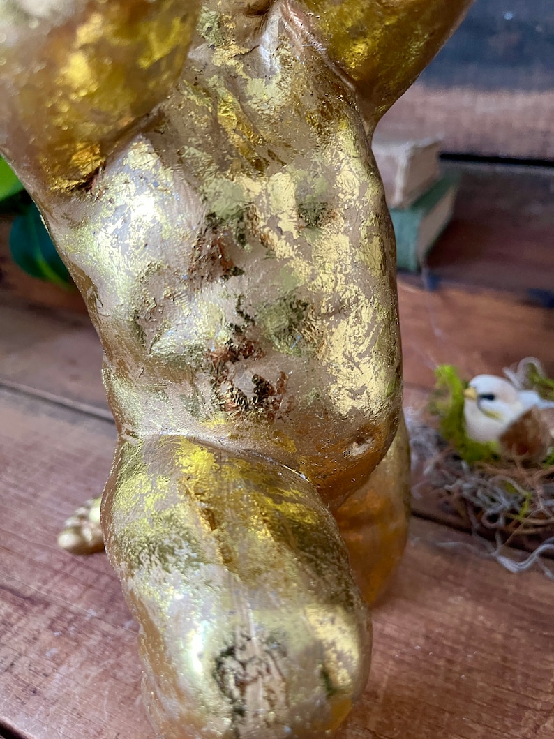 Puede incluir: Estatua de querub&iacute;n de color dorado con un acabado desgastado. La estatua est&aacute; en posici&oacute;n de pie con una pierna levantada y la otra doblada. El querub&iacute;n sostiene un objeto peque&ntilde;o en sus manos.