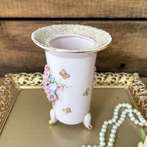 Peut inclure: Un vase en porcelaine blanche avec un bord dor&eacute; et un motif floral. Le vase a trois pieds et est d&eacute;cor&eacute; de fleurs roses et bleues et de papillons dor&eacute;s.