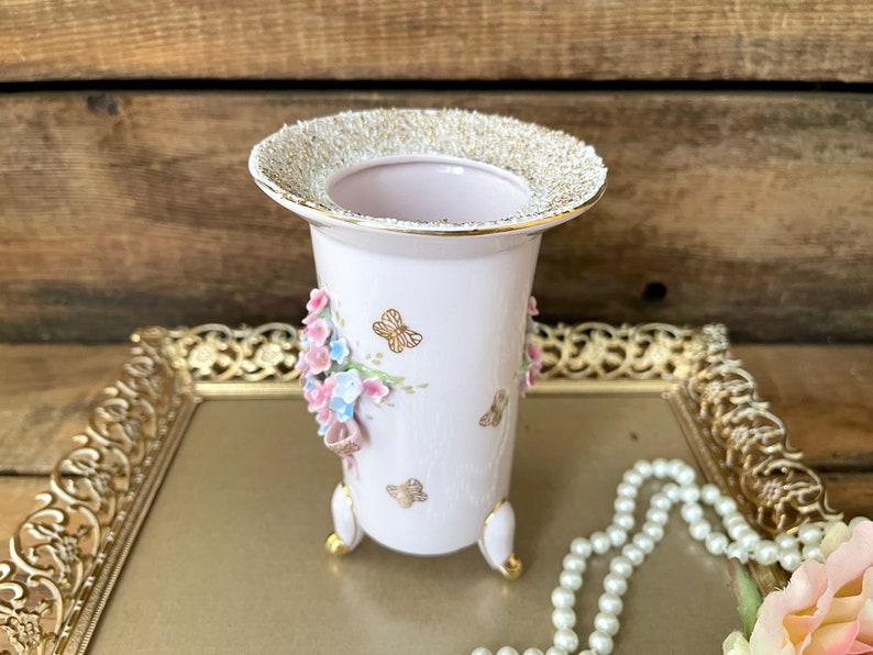 Peut inclure: Un vase en porcelaine blanche avec un bord dor&eacute; et un motif floral. Le vase a trois pieds et est d&eacute;cor&eacute; de fleurs roses et bleues et de papillons dor&eacute;s.