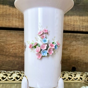 Peut inclure: Un vase en porcelaine blanche avec un bord dor&eacute; et trois pieds dor&eacute;s. Le vase est d&eacute;cor&eacute; d'un bouquet de fleurs roses et bleues.