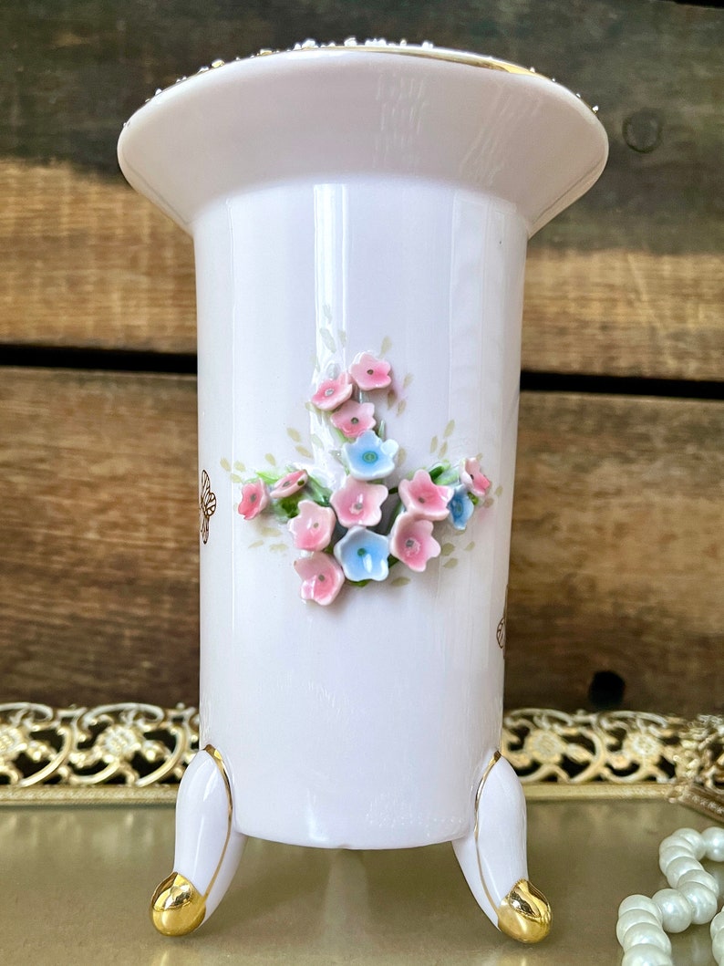 Peut inclure: Un vase en porcelaine blanche avec un bord dor&eacute; et trois pieds dor&eacute;s. Le vase est d&eacute;cor&eacute; d'un bouquet de fleurs roses et bleues.