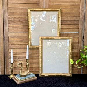 May include: Two rectangular gold picture frames with a bamboo-style border, two brass candlesticks with white taper candles, a brass swan figurine, and a stack of three books with green and brown covers arranged on a dark surface.