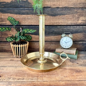 May include: A brass candle holder with a round base and a handle. The candle holder is on a wooden surface.