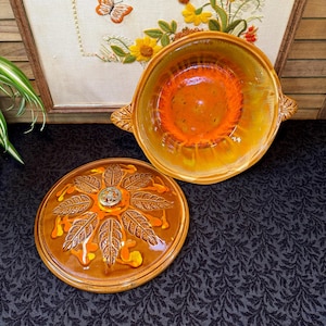 May include: Vintage ceramic casserole dish with lid. The dish is a warm orange and yellow with a glossy finish. The lid features a leaf design and a silver-colored knob. The dish has two handles and is open, revealing the interior.