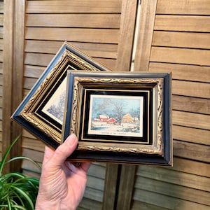 May include: Two framed winter landscape prints. The frames are black with gold trim. One print shows a snowy scene with red buildings. The other print has a black and white image. The text "WINTER IN THE COUNTRY" is visible.