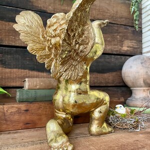 Puede incluir: Una estatua de querub&iacute;n dorada con las alas extendidas, arrodillada sobre una superficie de madera. La estatua tiene un acabado desgastado, lo que le da un aspecto antiguo.
