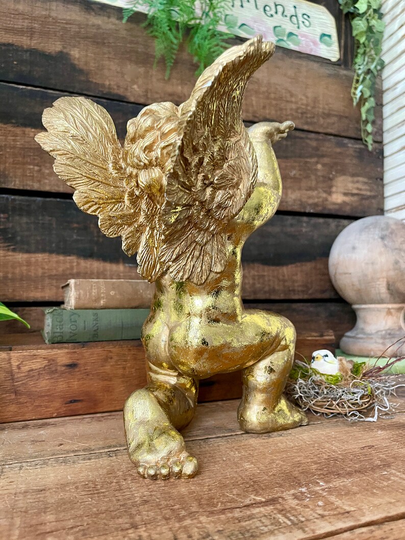Puede incluir: Una estatua de querub&iacute;n dorada con las alas extendidas, arrodillada sobre una superficie de madera. La estatua tiene un acabado desgastado, lo que le da un aspecto antiguo.