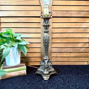 May include: An antique-style table lamp with a bronze finish. The lamp features a detailed, ornate base and a decorative column. A small potted plant and books are visible in the background. The lamp is designed to hold a light bulb.
