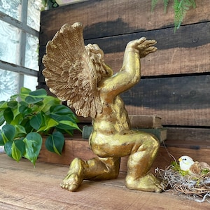 Puede incluir: Una estatua de querub&iacute;n dorada con los brazos y las alas extendidos, arrodillada sobre una superficie de madera. La estatua est&aacute; rodeada de vegetaci&oacute;n y un peque&ntilde;o nido de p&aacute;jaro.