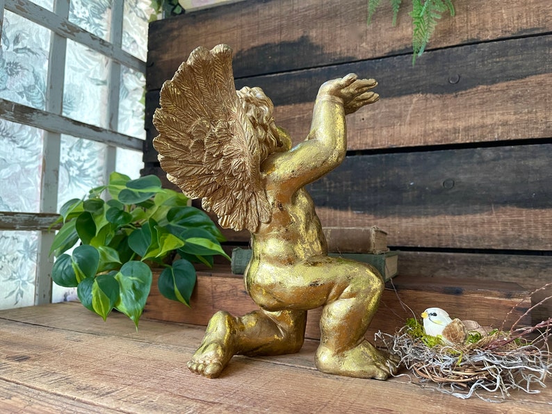 Puede incluir: Una estatua de querub&iacute;n dorada con los brazos y las alas extendidos, arrodillada sobre una superficie de madera. La estatua est&aacute; rodeada de vegetaci&oacute;n y un peque&ntilde;o nido de p&aacute;jaro.