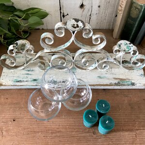 Op de afbeelding: Witte metalen kaarsenhouder met een verweerde afwerking, met een gerold ontwerp. De houder is gemonteerd op een houten basis met een vergelijkbare verweerde afwerking. Drie heldere glazen theelichthouders en drie tealgroene theelichtjes zijn ook inbegrepen.