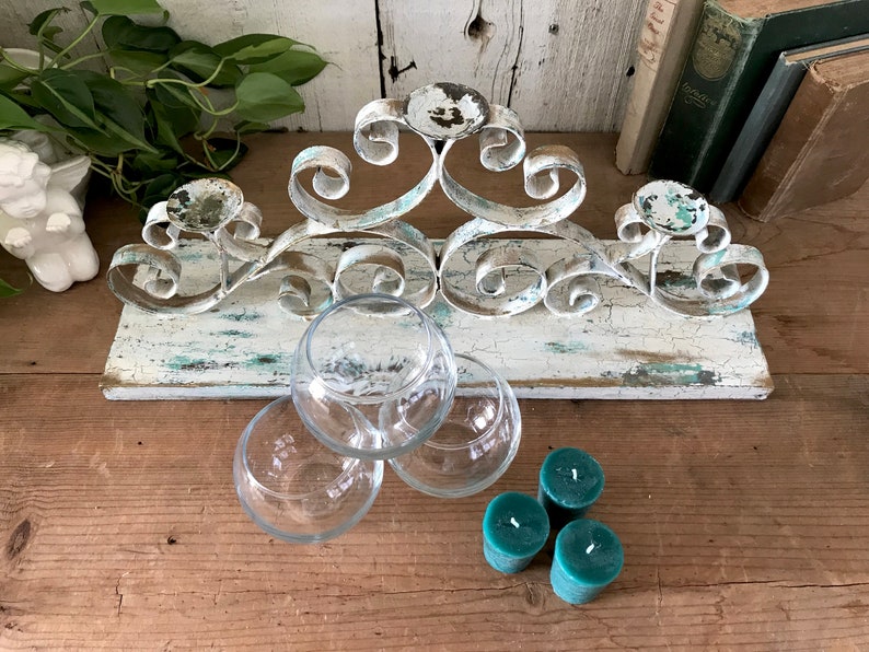 Op de afbeelding: Witte metalen kaarsenhouder met een verweerde afwerking, met een gerold ontwerp. De houder is gemonteerd op een houten basis met een vergelijkbare verweerde afwerking. Drie heldere glazen theelichthouders en drie tealgroene theelichtjes zijn ook inbegrepen.