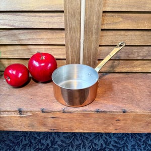 May include: A small copper saucepan with a brass handle sits on a wooden surface, next to two shiny red apples. The pan has a silver interior and is empty. The background is a wooden shutter.