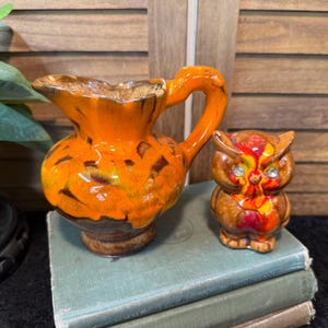 May include: An orange ceramic pitcher with a handle and a brown base sits next to a small owl figurine. The pitcher has a textured surface with dark brown accents. The owl figurine has red, yellow, and brown details.