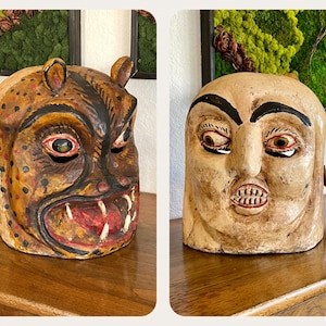 May include: Two hand-carved wooden masks with painted details. The mask on the left is a fierce animal with a spotted pattern, while the mask on the right is a human face with a frowning expression.
