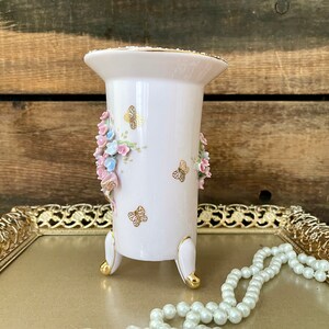 Peut inclure: Un vase en porcelaine blanche avec des accents dor&eacute;s et un motif floral compos&eacute; de fleurs roses et bleues et de papillons. Le vase a trois pieds dor&eacute;s et est pos&eacute; sur un plateau dor&eacute; avec un collier de perles.